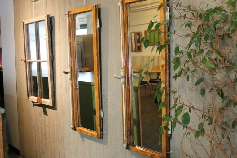 An einer grauen Wand hängen alte Fensterrahmen, deren Fensterglas durch Spiegel ersetzt wurde.